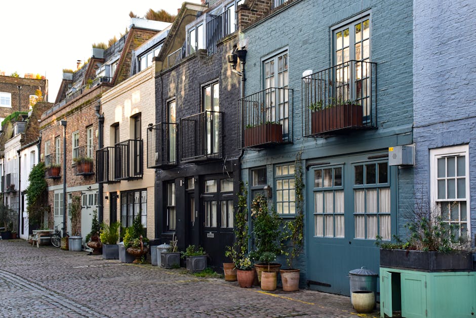 A row of narrow, multi-storey residential buildings situated along a cobblestone street. The buildings feature various exterior finishes, including brick and painted surfaces in shades of light blue, beige, grey, and black. Several of the houses have small balconies with black metal railings, some of which are adorned with potted plants. In front of the buildings, there are numerous potted plants and small shrubs placed directly on the pavement and on window sills, giving a touch of greenery to the scene. A white wooden bench sits on the sidewalk near one of the houses, and a bicycle leans against a wall in the background. The environment appears to be urban, with a peaceful, residential atmosphere, and the scene is illuminated by natural daylight, highlighting the textures of the brickwork and plant foliage. Waste Clearance Soho occasionally handles tasks involving the collection and disposal of household waste, as illustrated by the clean, uncluttered appearance of the front yards, which have been tidily maintained and cleared of bulk rubbish, exemplifying professional private waste handling in a city setting.