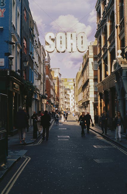 A bustling street scene in Soho, London, featuring a large, illuminated sign spelling 'SOHO' suspended overhead between narrow, multi-storey buildings on either side of the street. The buildings exhibit a mix of architectural styles, with some having detailed facades, decorative railings, and large windows. The street is populated with pedestrians walking in both directions, some dressed in dark clothing, and others with lighter or patterned attire, reflecting a lively urban atmosphere. The pavement shows a combination of traditional bricks and painted road markings. In the background, the street extends into further buildings with varying heights, and the sky above is partly cloudy with patches of blue. The scene exemplifies an active central London district, often associated with independent shops, restaurants, and cultural venues, resonating with alternative waste handling services such as private rubbish collection and on-site clearance offered by Waste Clearance Soho, visible through the context of urban waste management and street activity.
