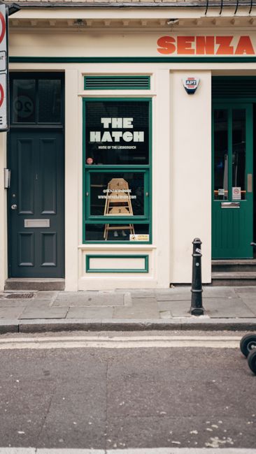 The image depicts the exterior front of a small commercial building with a central glass window and door framed in teal, situated between a solid dark gray door on the left and a teal-painted shopfront on the right. The window displays a sign with the phrase 'THE HATEL' in bold white letters, and beneath it, smaller text indicates the business's location in the Lighthouse. A wooden stool with a step ladder resting on top is visible inside the shop through the window. The building's facade features a cream-colored wall with teal trim and decorative paneling underneath the window. Above the shop, part of a red and orange sign reads 'SENZA,' with only a partial view of the branding. A small security alarm is mounted on the upper right corner of the wall, and a black bollard is positioned on the sidewalk in front of the building. The environment suggests a city street setting with a narrow pavement and street markings visible. Inside the scene, there are no visible waste or rubbish items, indicating a tidy storefront in an urban area that may utilize private waste collection services, consistent with the context of alternative rubbish removal options in Soho Square businesses.