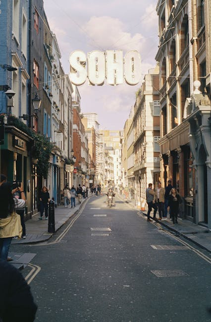 A busy street scene in Soho, featuring a narrow road lined with multi-story buildings on both sides that display a mix of modern and traditional architecture with brick facades, large windows, and decorative awnings. Pedestrians are walking along the pavement, some in groups and others alone, while a cyclist rides down the centre of the street. In the background, a large white sign spelling 'SOHO' in bold, capital letters is suspended above the street, with a cloudy sky overhead. The street surface shows typical urban markings, and there are details like street lamps, bollards, and small shopfronts visible along the sidewalk. The scene captures the vibrant, urban atmosphere of a commercial and entertainment district, where private disposal and independent waste collection services like Waste Clearance Soho may operate to handle rubbish from local businesses and residents, supporting effective rubbish removal in the area.