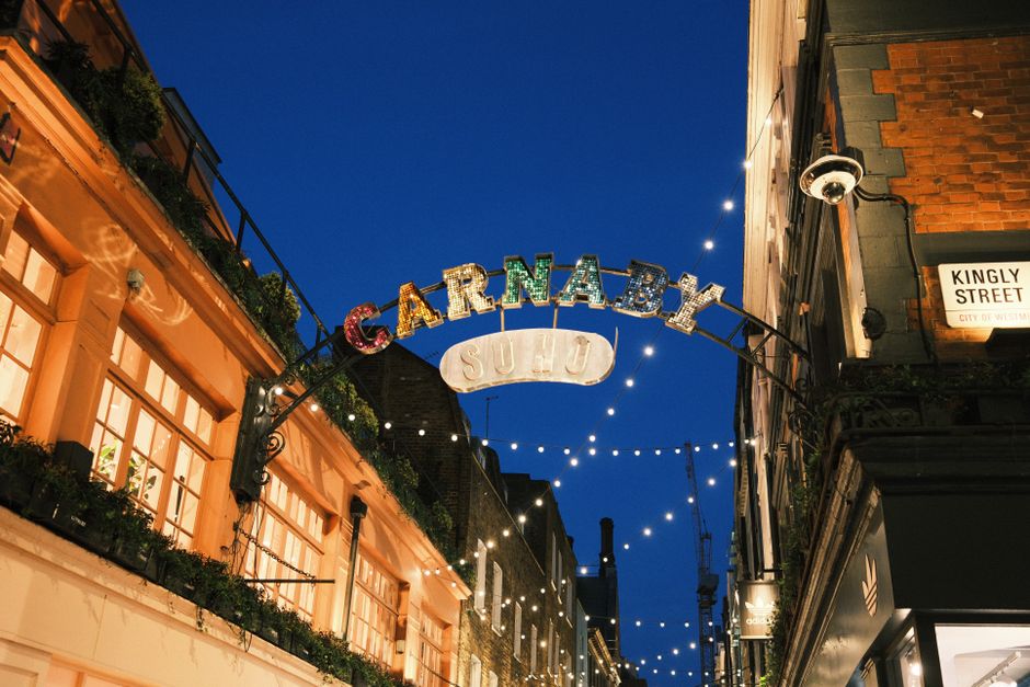 A nighttime street scene featuring an illuminated sign spelling 'CARNABY' in multicoloured, vintage-style letters, arching over a narrow pedestrian alley. The sign is mounted on a black metal frame attached to brick and stucco buildings on either side of the street. To the left, the building has warm, orange-toned exterior walls with large windows, some of which are decorated with plants and string lights. On the right, part of a dark-colored building with a street sign indicating 'Kingly Street' is visible, along with architectural details like brickwork and a security camera mounted near the corner. The sky overhead is a deep blue, contrasting with the bright, festive string lights hanging across the street, creating a lively and welcoming atmosphere associated with vibrant nightlife and commercial activity in areas like Carnaby Street Soho. This scene reflects an urban environment prepared for evening visitors, with a focus on the distinct signage typical of independent retail and entertainment venues, relevant to the context of private waste handling or rubbish clearance services like those provided by Waste Clearance Soho.