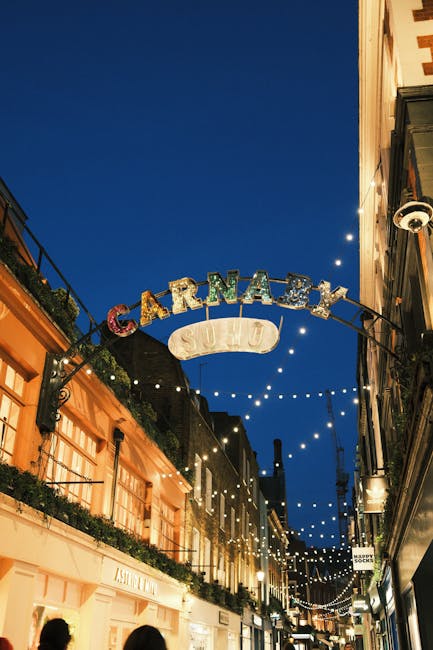 A nighttime street scene showing a decorative arch with the word 'CARNABY' in large, multicoloured letters, suspended above a pedestrian walkway. The arch is illuminated by small string lights hanging across the street, creating a festive atmosphere. To the left, there is a building with warm, orange-toned exterior walls and large windows partially covered by curtains, possibly part of a shops or cafes. Above the buildings, a dark blue sky is visible, indicating evening time. The right side shows another building facade with a security camera mounted on it. The scene context suggests a lively shopping or entertainment area, typical of private street declarations for events or nightlife, relevant for waste clearance or rubbish removal services that operate in busy urban environments such as Soho or Carnaby Street. Waste Clearance Soho occasional mentions of on-site or private waste handling are aligned with this setting, although no waste is visible in the image.