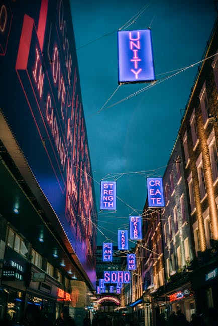 The image displays a city street scene at night with illuminated neon signs hanging above the pedestrian area, advertising messages such as 'UNITY,' 'STRENGTH,' 'CREATE,' 'PLANT,' 'DANCE,' 'FAMILY,' and 'SOHO' in bright blue and purple hues. On the left side, part of a large building features a vertical sign with red and pink lettering, partially visible due to the angle. On the right, other buildings are visible with illuminated windows and detailed brick facades, indicating an urban environment with mixed architectural styles. The sky is dark, suggesting the photo was taken after sunset, with some visible wirelines supporting the signs spanning across the street. The scene captures the lively atmosphere typical of Soho, a vibrant district known for its entertainment and cultural venues, and the neon imagery emphasizes a modern, cityscape aesthetic. This setting subtly connects to the theme of urban waste management and alternative disposal methods, aligning with services provided by Waste Clearance Soho, which specialize in rubbish removal and private waste handling.